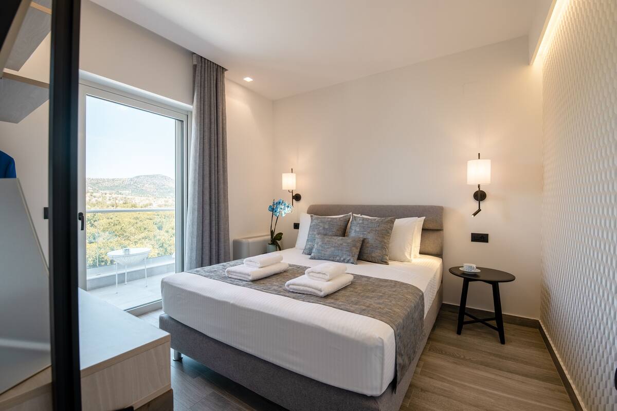A modern hotel room with a large bed, two bedside tables, a small side table, and a window with a view of the outdoors.