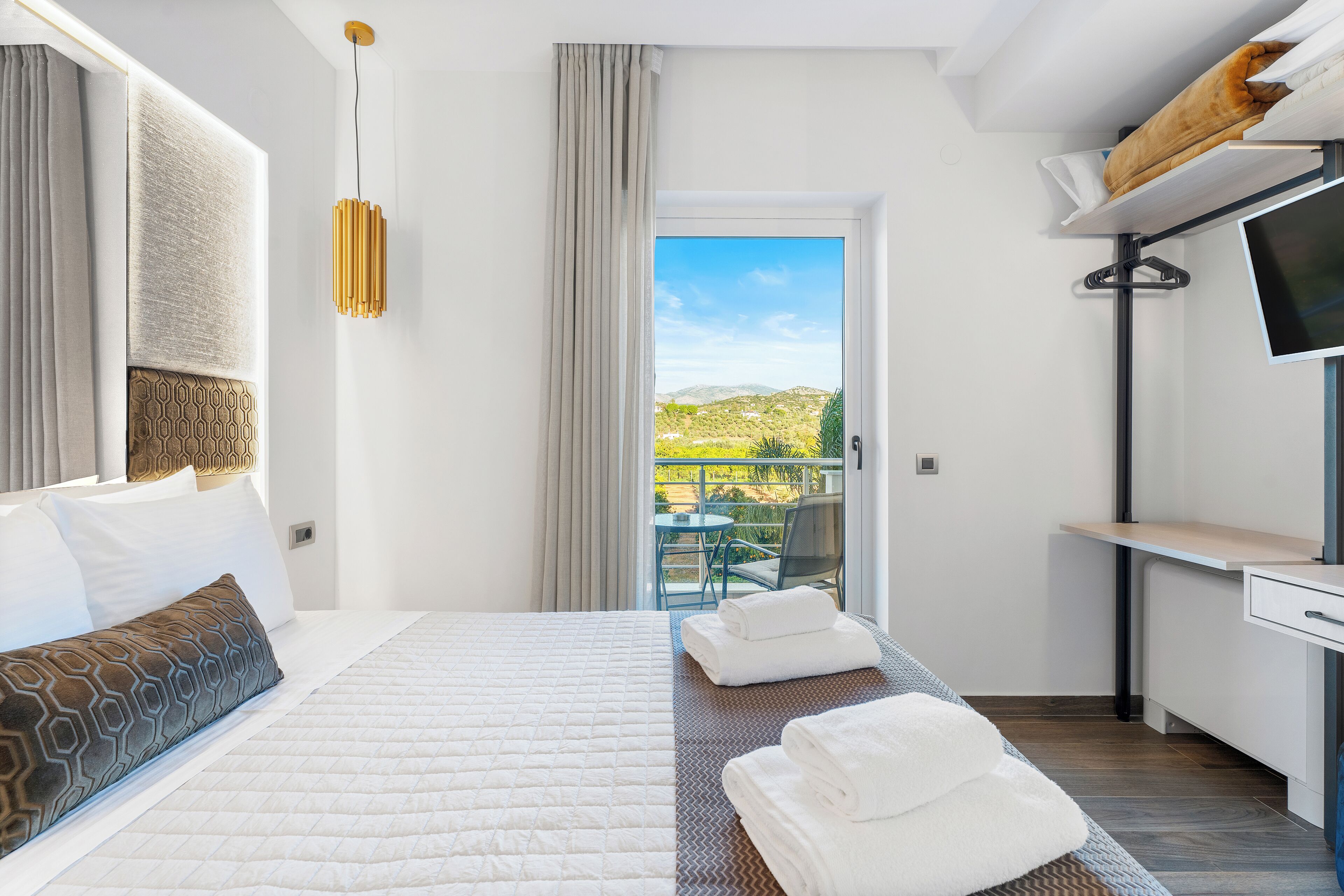 Una habitación de hotel moderna con una cama, un escritorio, un televisor y un balcón con vistas a las colinas.