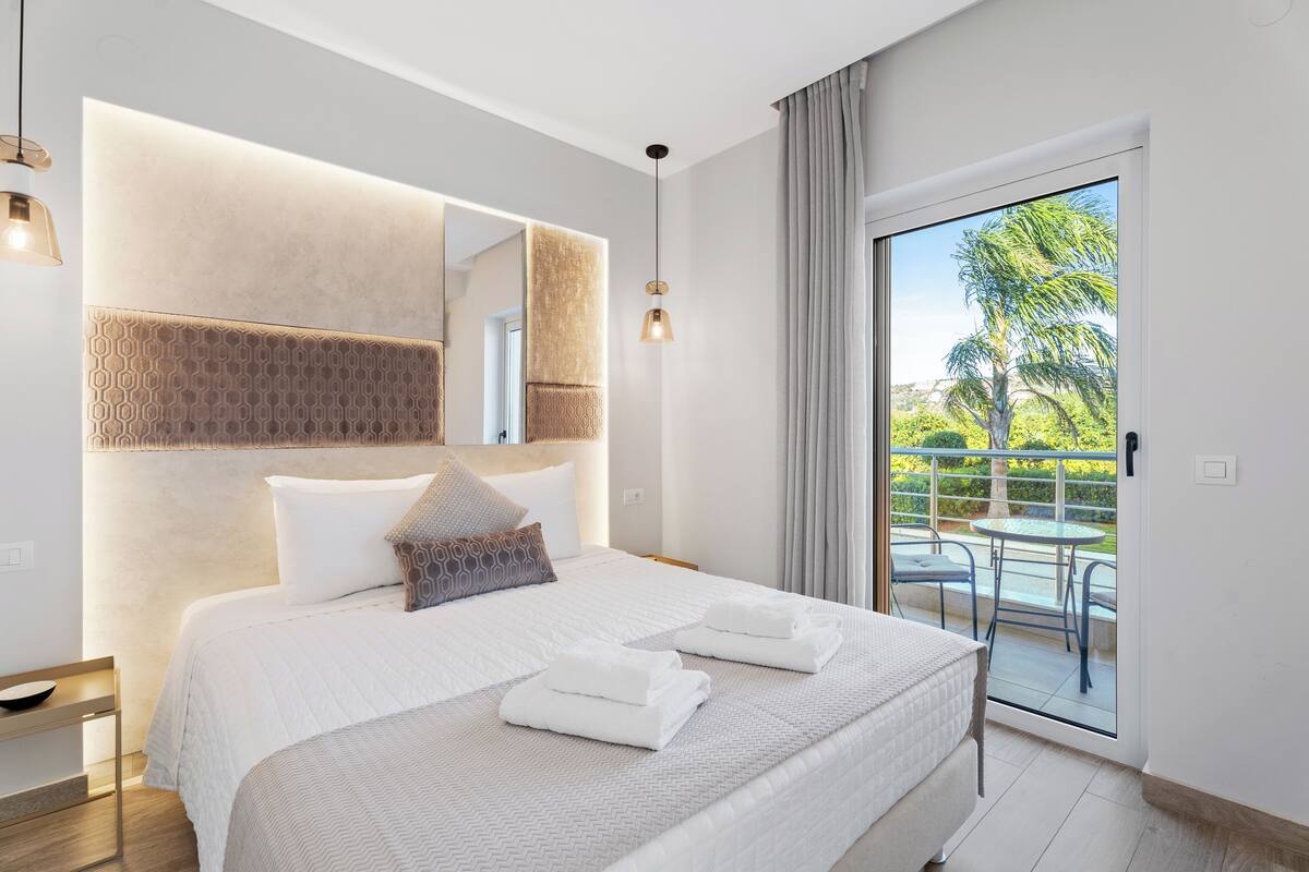 A modern hotel room with a large bed, a balcony with a view of palm trees, and a well-lit ceiling.