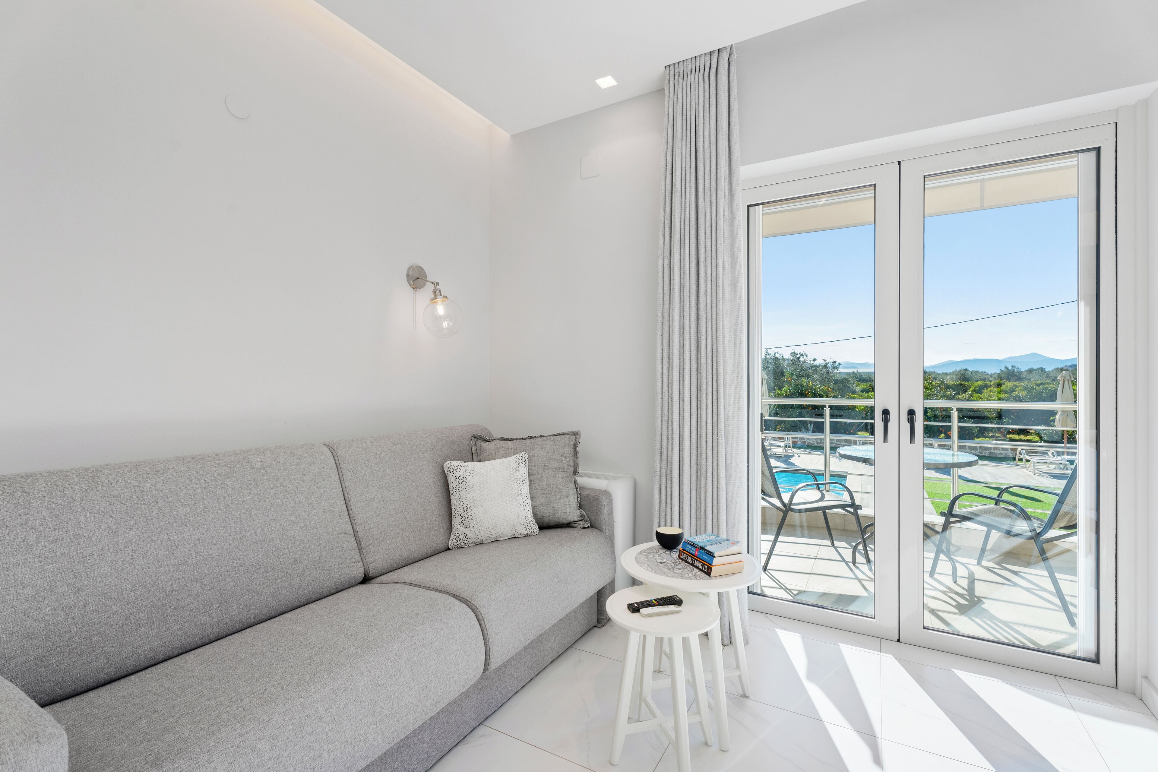 A modern living room with a grey sofa, a small white table, and a view of a pool and greenery through large sliding doors.