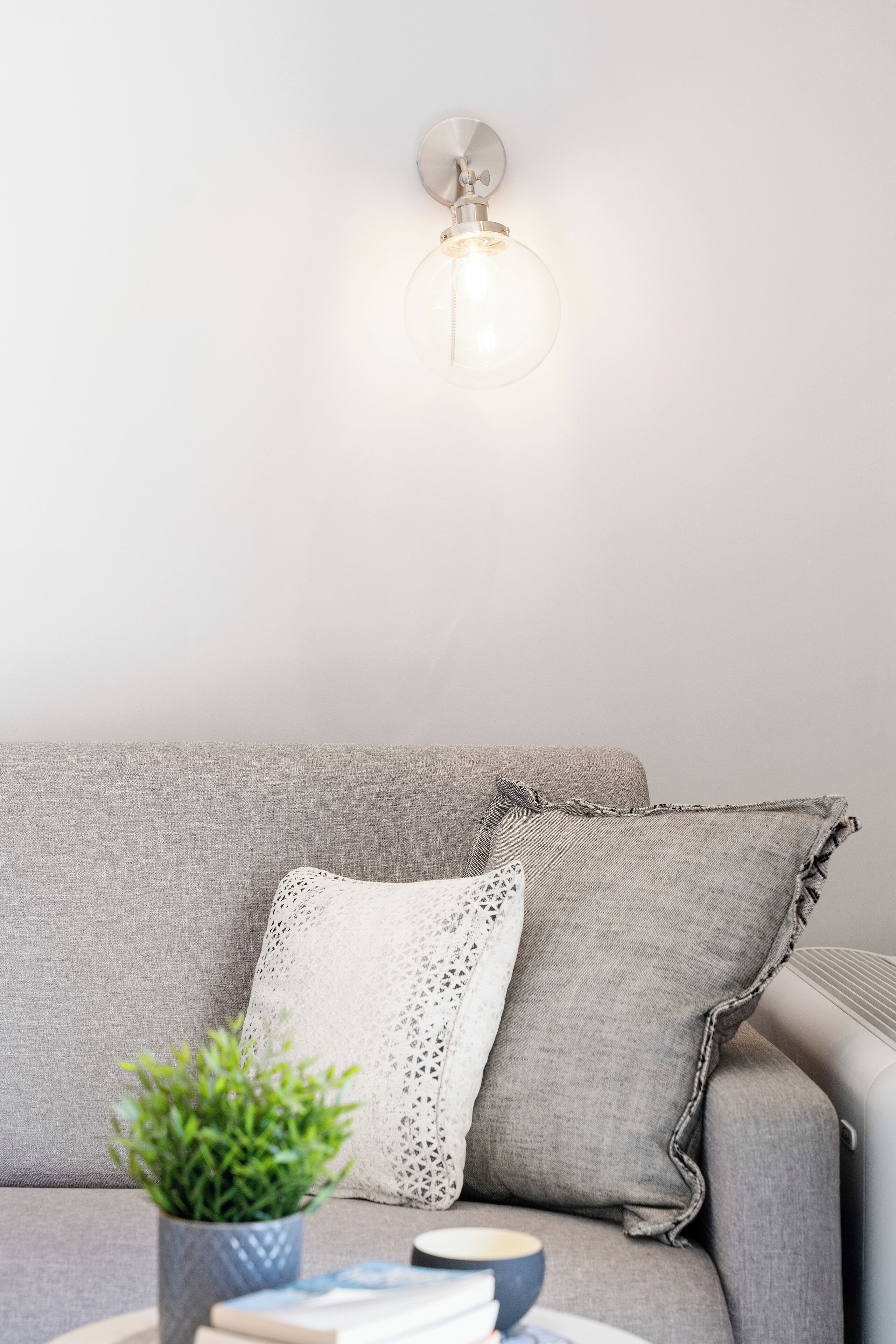 Un sofá gris con un cojín blanco de punto y un cojín gris con flecos. Una pequeña planta en maceta está sobre el sofá.