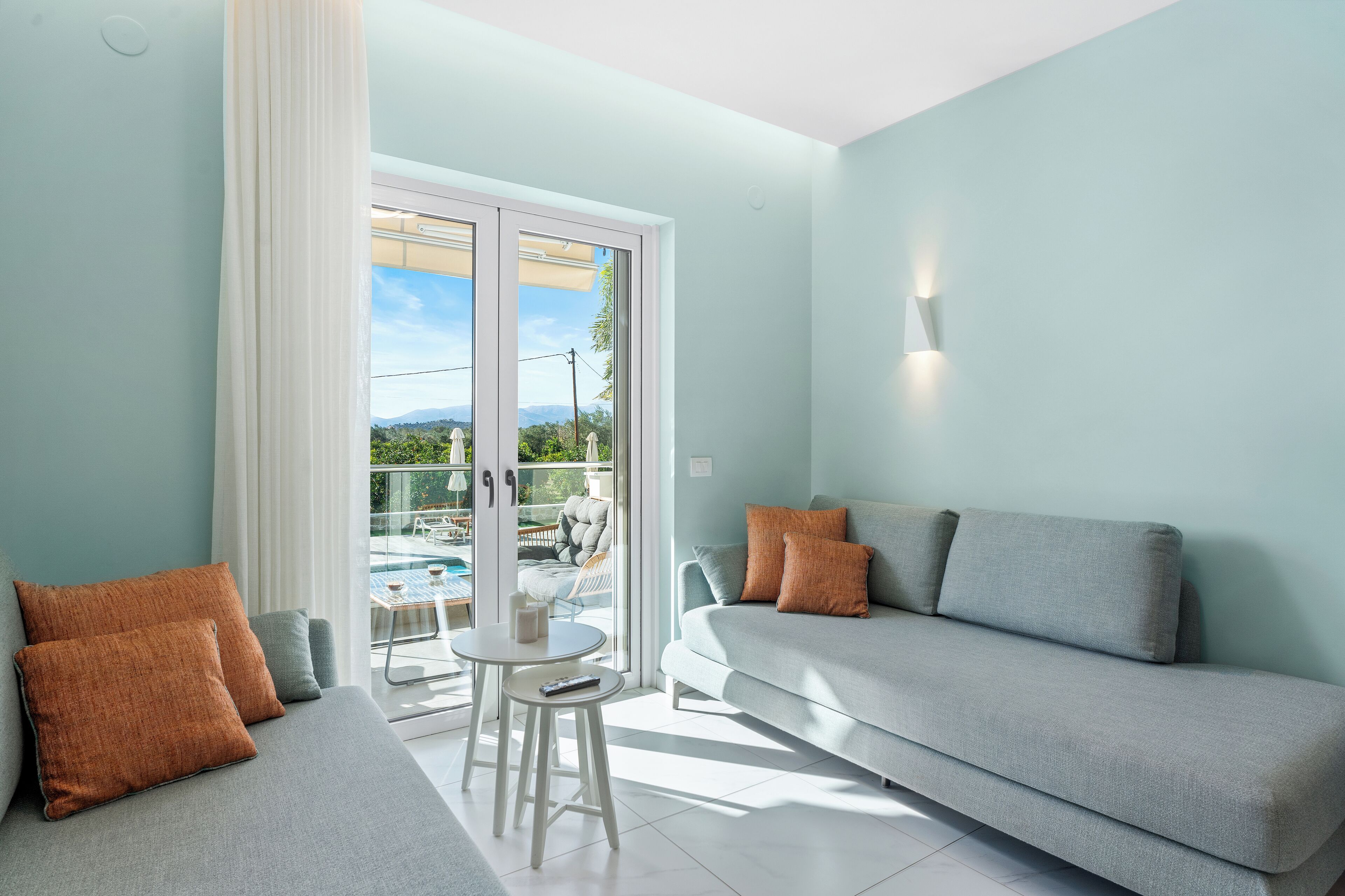 Un salón moderno con un sofá gris, dos cojines naranjas, una mesita blanca y un ventanal de vidrio que da a un balcón con vistas a una piscina.