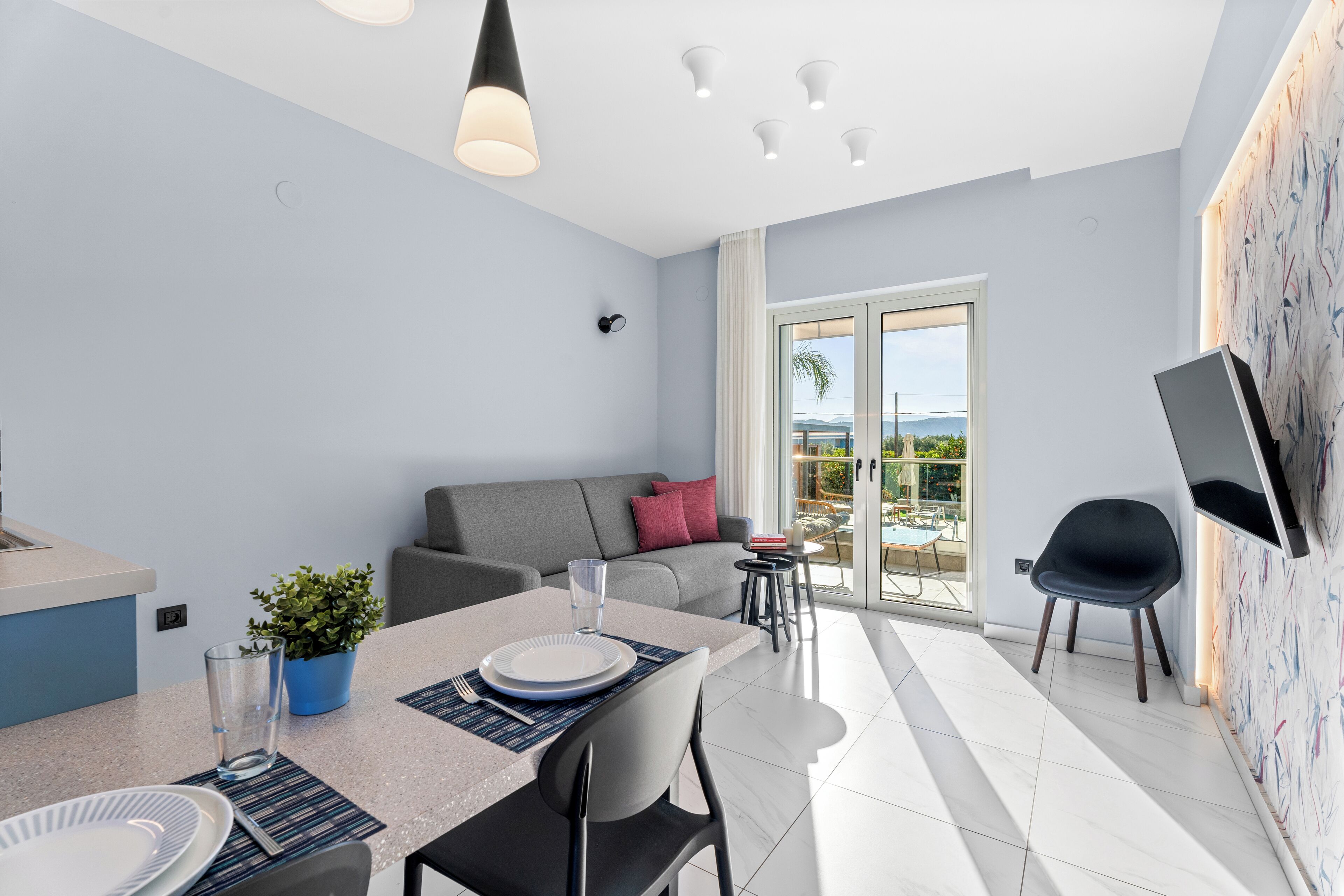 A modern living room with a dining area, a grey sofa, a TV mounted on the wall, and a balcony with a view.