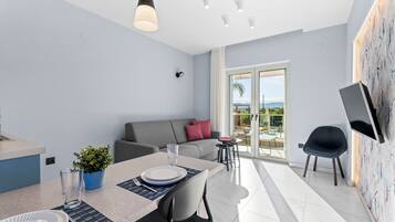 A modern living room with a dining area, a grey sofa, a TV mounted on the wall, and a balcony with a view.