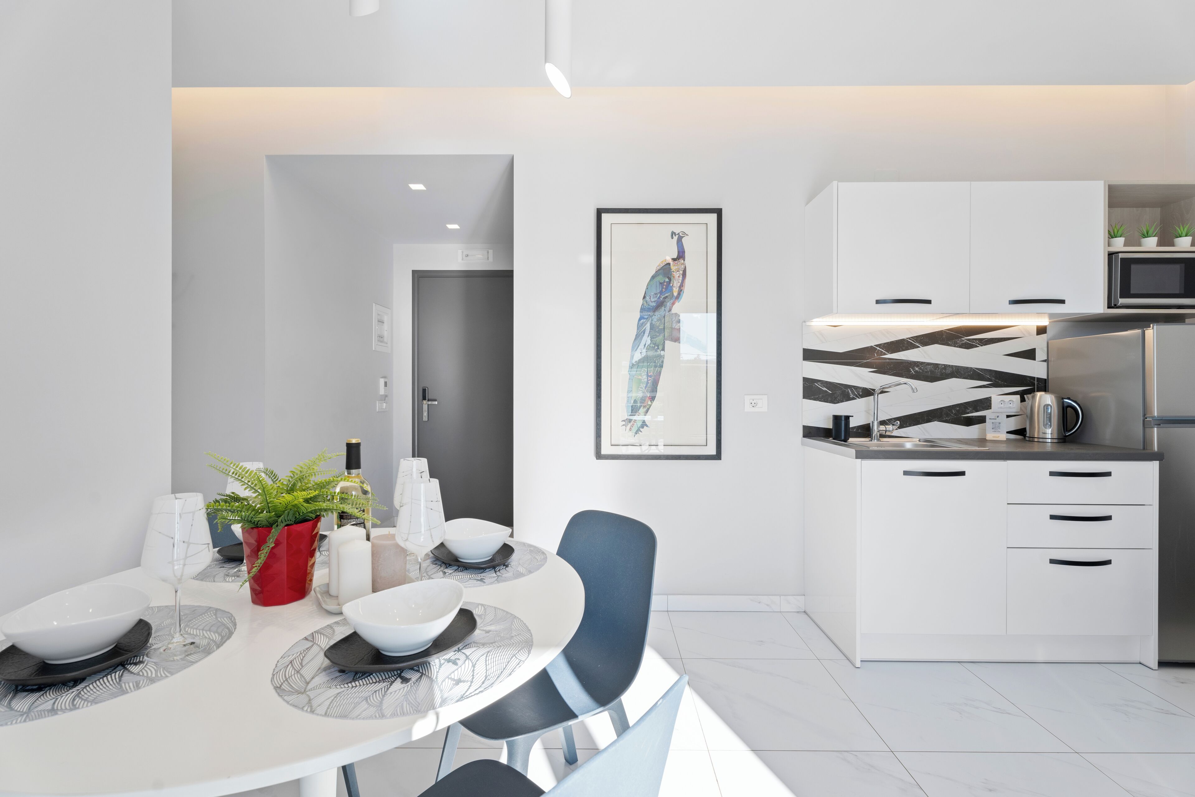 A modern kitchen with white cabinets, a microwave, and a refrigerator. There is a dining table with chairs and a potted plant.