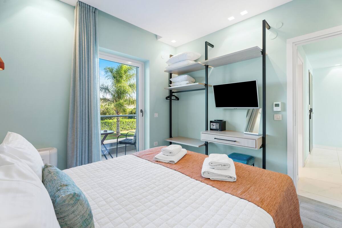 A hotel room with a bed, a TV, a desk, and a balcony with a view of greenery.