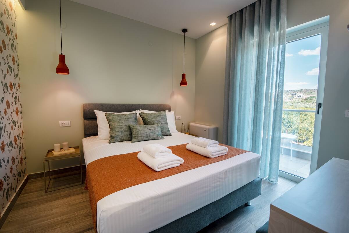 A bedroom with a bed, two bedside tables, a nightstand, a window with curtains, and a view of the outdoors.