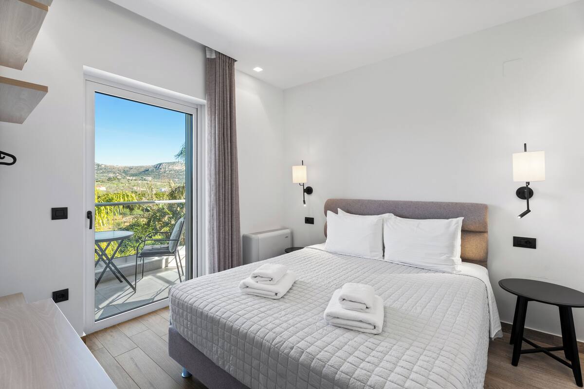 A modern hotel room with a large bed, two bedside tables, a small table, and a balcony with a view of the countryside.