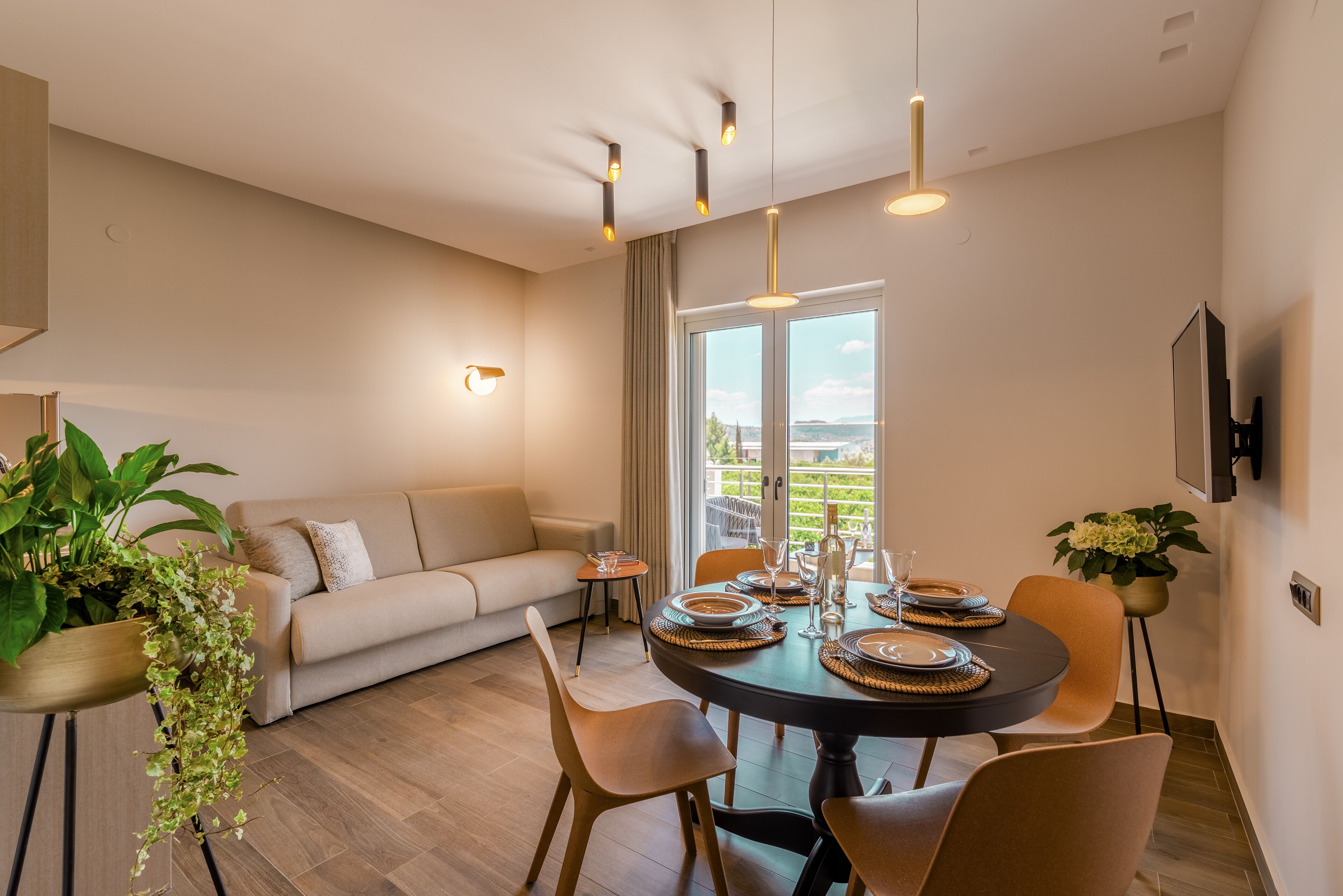 Un comedor con una mesa redonda preparada para cuatro, rodeada de sillas. Un sofá está ubicado contra la pared, y un ventanal ofrece vistas al exterior.