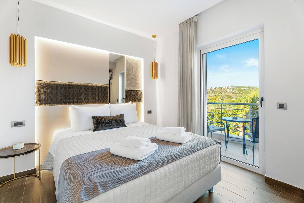 A modern hotel room with a large bed, a small table, and a balcony with a table and chairs.