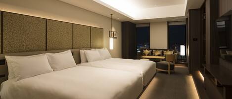 A modern hotel room with a large bed, a seating area with a sofa and armchairs, a TV, and a city view through the windows.