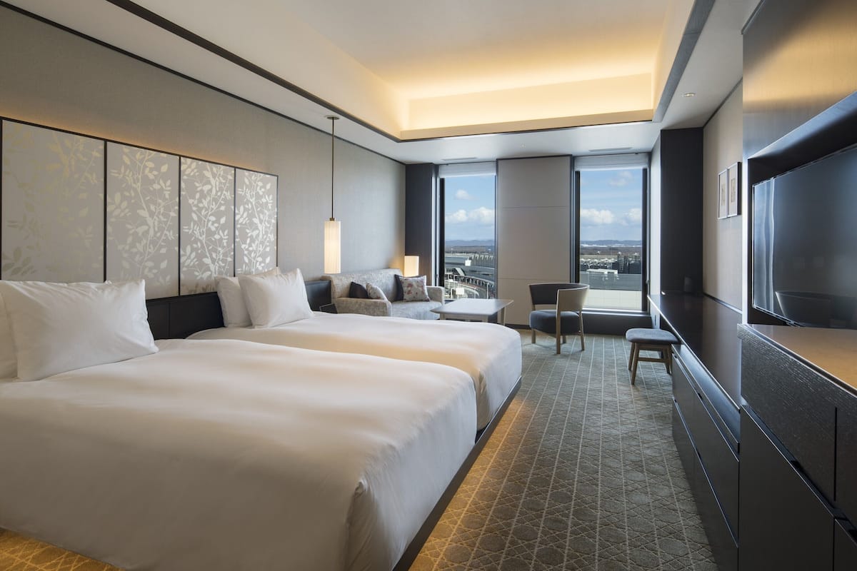 Ein Hotelzimmer mit zwei Betten, einer Sitzecke, einem Fernseher und einem großen Fenster mit Blick auf die Stadt.