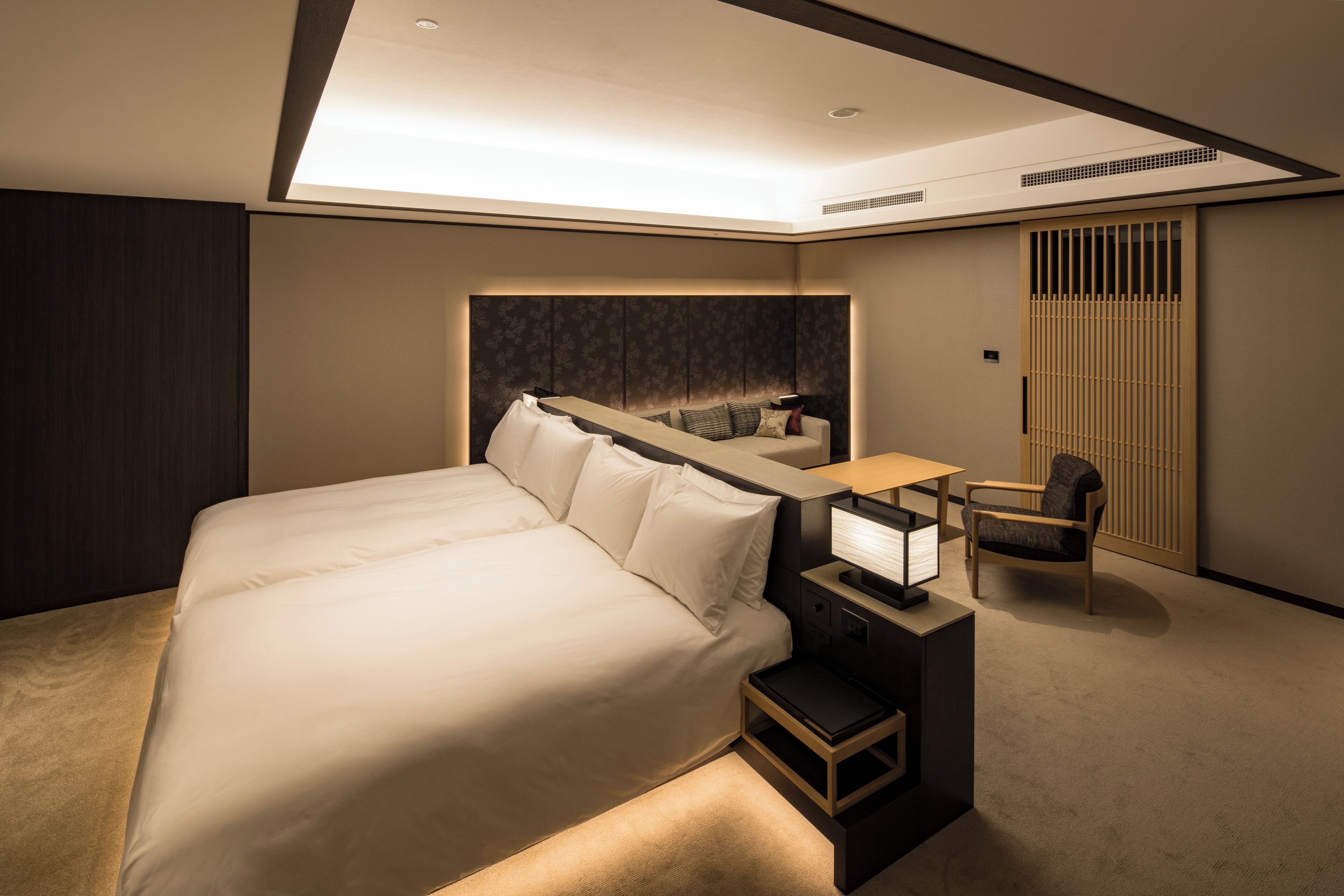 A modern hotel room with a large bed, a desk with a lamp, a chair, and a wardrobe.