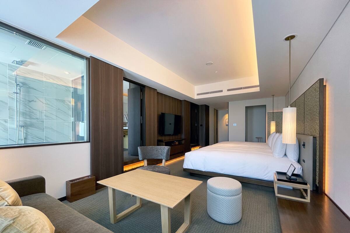 Ein modernes Hotelzimmer mit einem großen Bett, einer Couch, einem kleinen Tisch, einem Fernseher und einem großen Fenster mit Blick auf die Stadt.