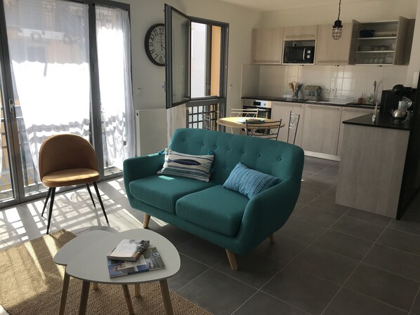 A modern living room with a teal sofa, a small white table, and a chair. A kitchen with a microwave and cabinets is visible in the background.