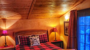 A wooden bed with a checkered bedspread and a pillow with 'LET THE ADVENTURE BEGIN' text, flanked by two bedside tables with lamps, in a room with wooden ceiling beams and a window with curtains.