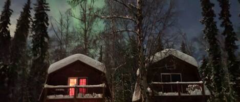 A cabin with a snow-covered roof and a lit window, surrounded by trees and snow.