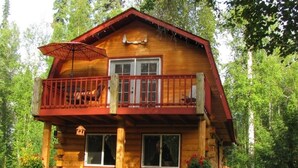 A two-story log cabin with a balcony, surrounded by lush greenery.