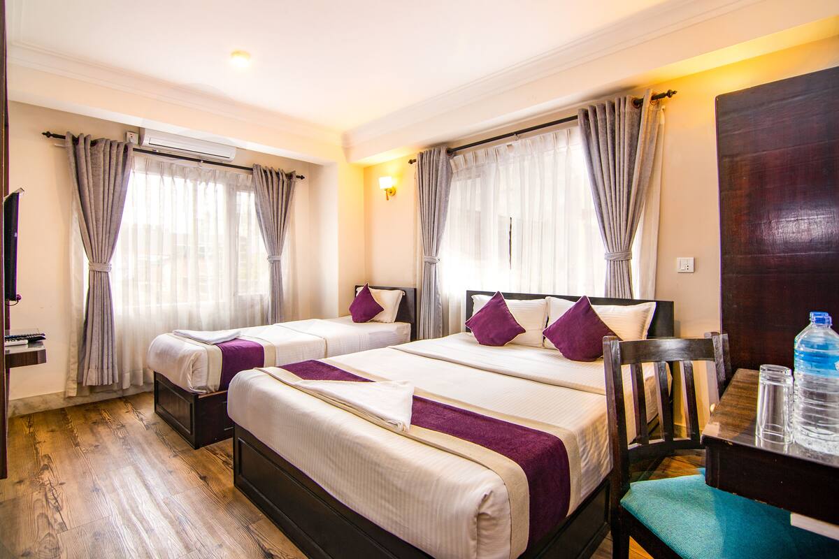 A hotel room with two beds, a wooden floor, a desk with a chair, and a large window with curtains.