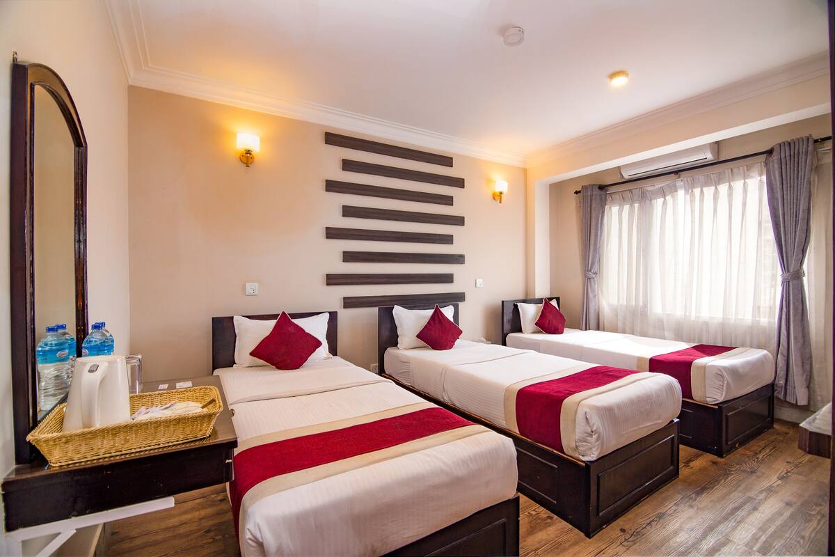 A hotel room with three beds, a large mirror, a wall-mounted light, and a window with curtains.