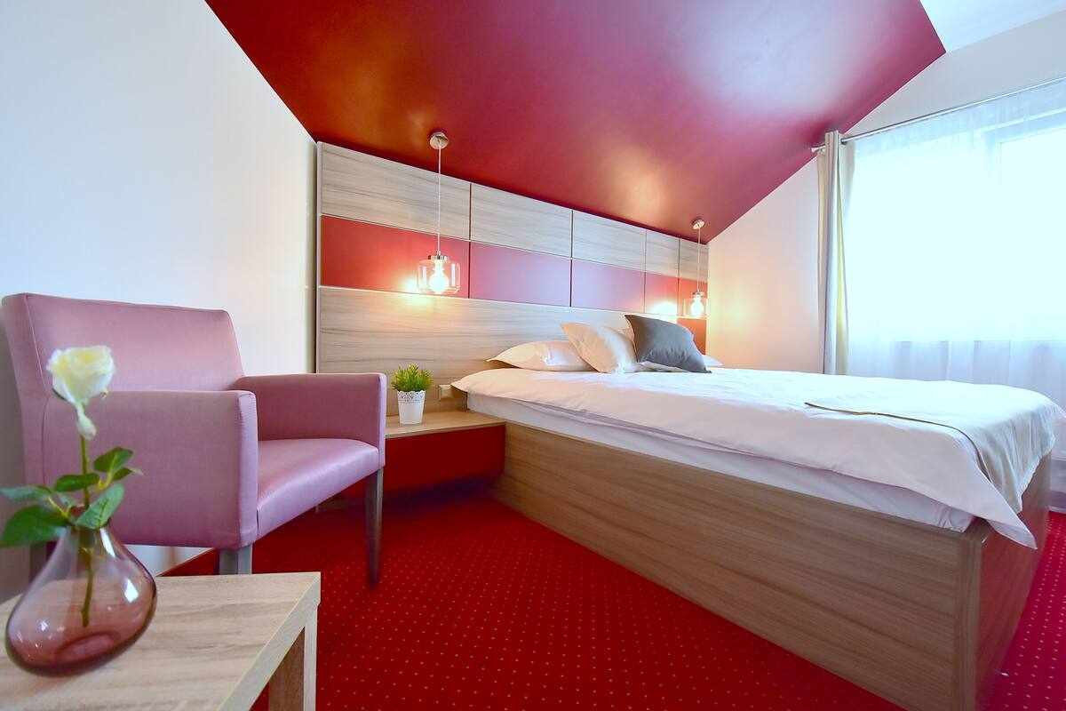 A hotel room with a bed, a chair, a small table, and a vase with a flower.