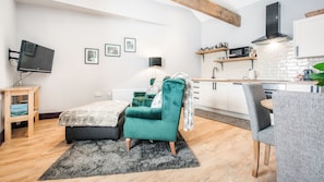 A modern living room with a teal sofa, a small table, and a TV on a wooden stand. The room has a kitchen with white cabinets and a black microwave. - Village Farm Cottages.  (Driffield)
