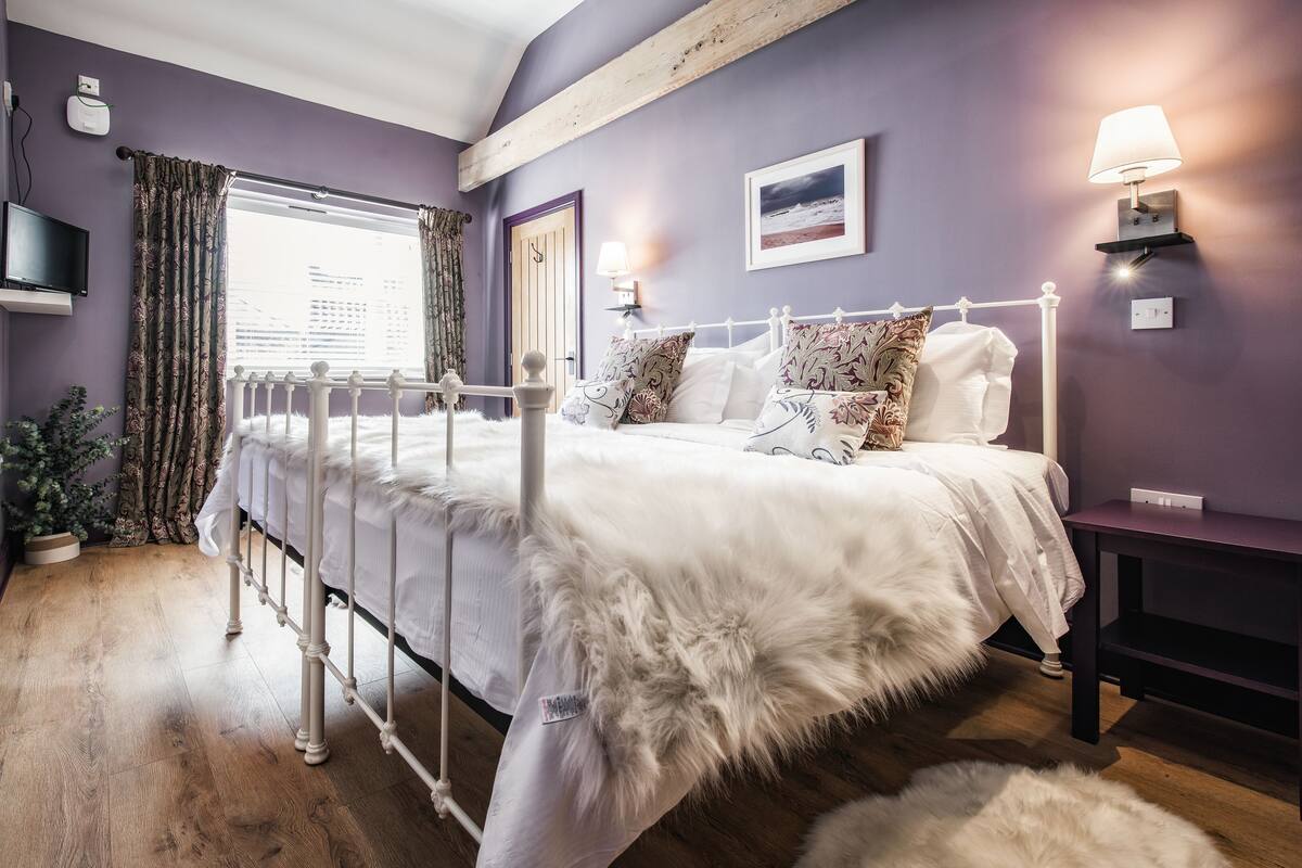 A bedroom with a white bed, purple walls, a window with curtains, a bedside lamp, and a framed picture on the wall.