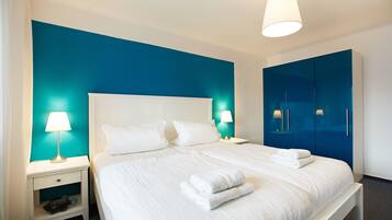 A neatly made bed with white linens and towels, a white nightstand with a lamp, and a blue accent wall.