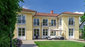 Ein zweistöckiges, gelbes Gebäude mit rotem Ziegeldach, großen Fenstern und einer kleinen Terrasse mit Gartenmöbeln.