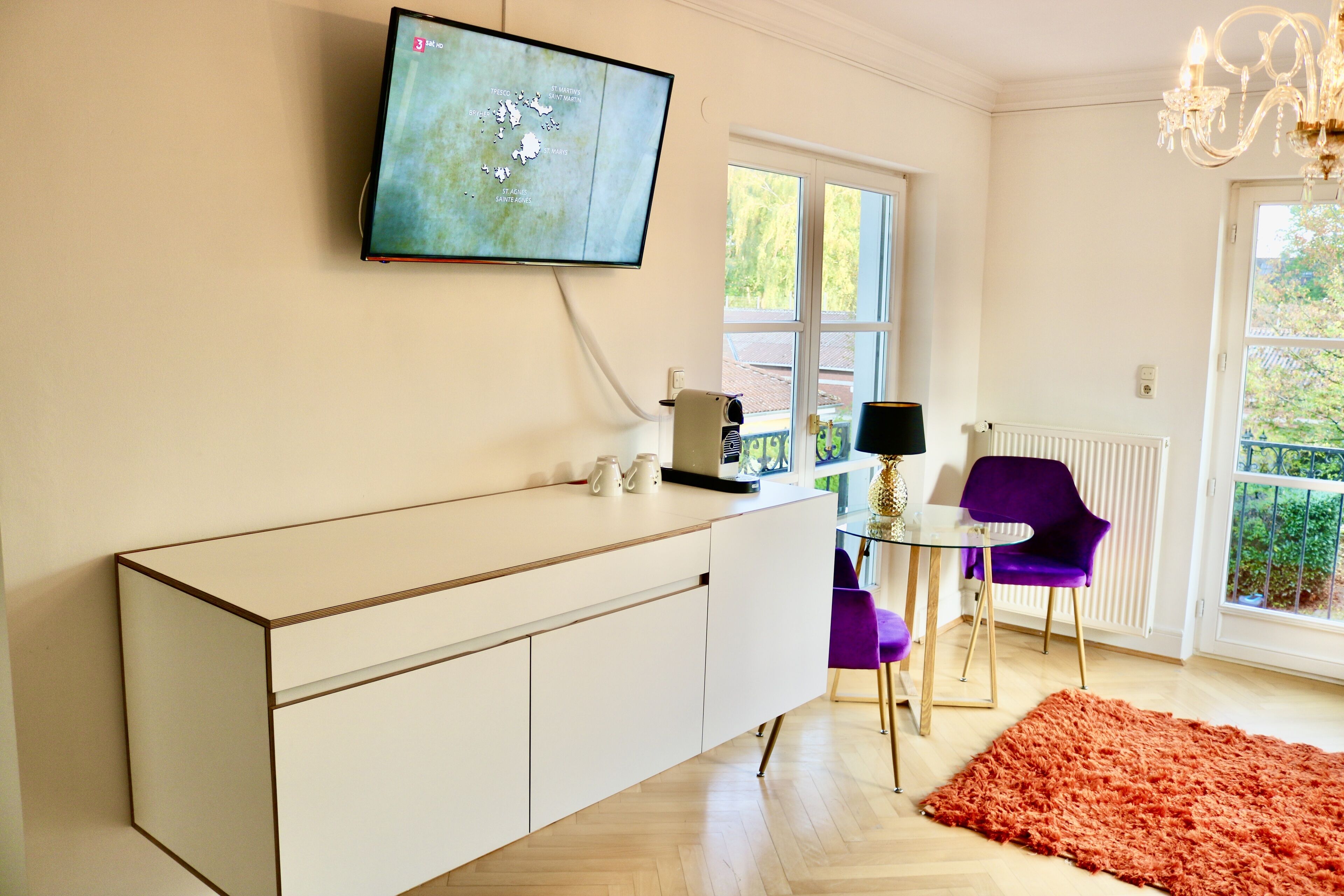 A modern living room with a flat-screen TV mounted on the wall, a white cabinet, a glass coffee table, purple chairs, and a large window with a view of greenery.