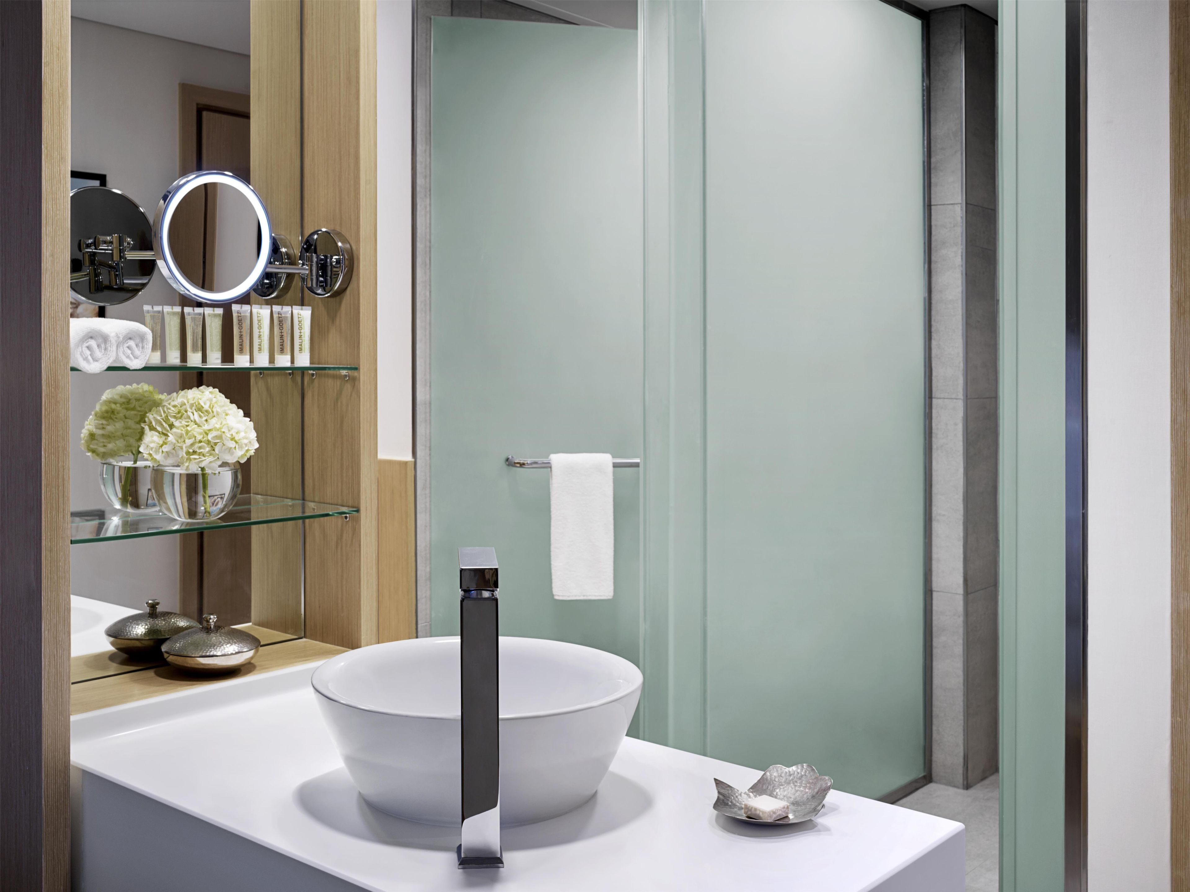 Ein modernes Badezimmer mit einem großen, weißen Waschbecken, einem Spiegel mit Beleuchtung, einem Handtuchhalter und einer Duschkabine aus Glas.