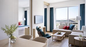 A modern hotel room with a large window, a sofa, a coffee table, and a dining area.