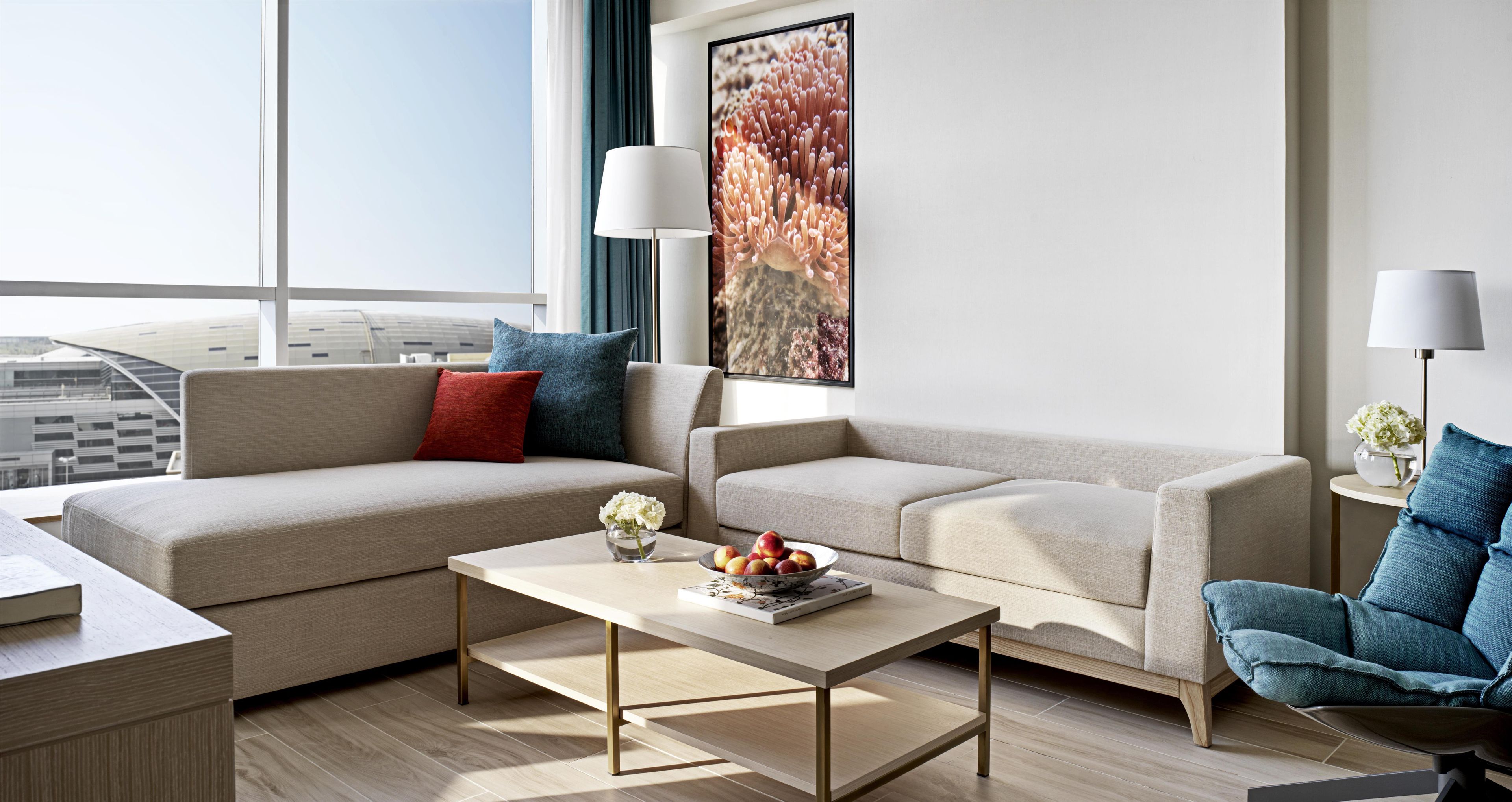 A modern living room with two sofas, a coffee table, and a chair. There is a window with a view of a building, and a painting on the wall.