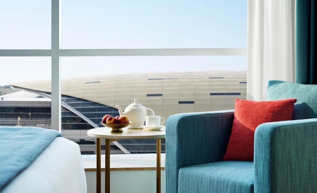 Ein Hotelzimmer mit Blick auf ein modernes Gebäude, ein kleiner Tisch mit Teekanne und Obst sowie ein blauer Sessel mit rotem Kissen.