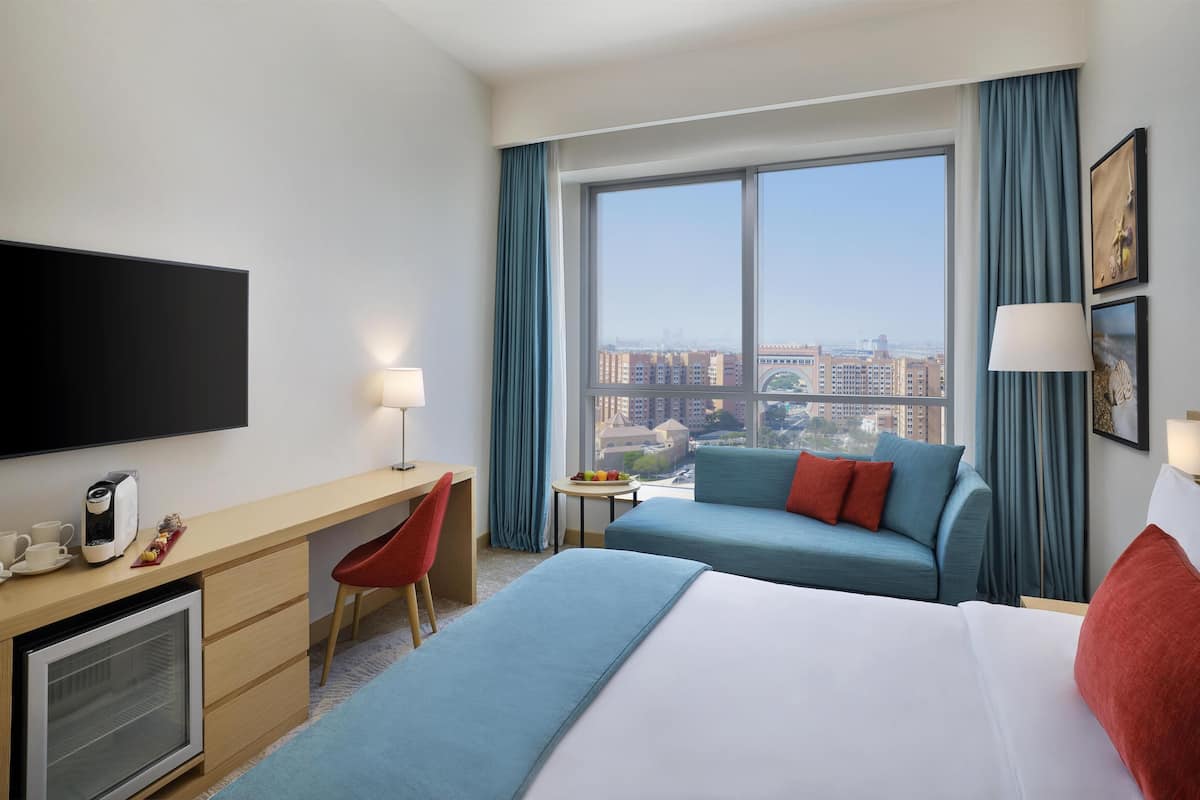 A hotel room with a large bed, a desk with a flat-screen TV, a small table with fruit, a sofa, and a view of the city through large windows.