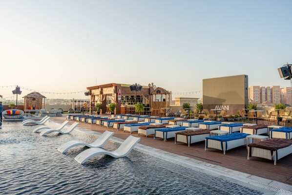 Un área de piscina en la azotea con sillones y mesas, un edificio con el letrero 'AVANI', y un paisaje urbano al fondo.