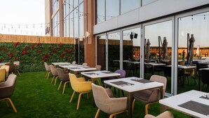 An outdoor seating area with tables and chairs, a green lawn, and a building with large windows. - Avani Ibn Battuta Dubai Hotel (Dubai)