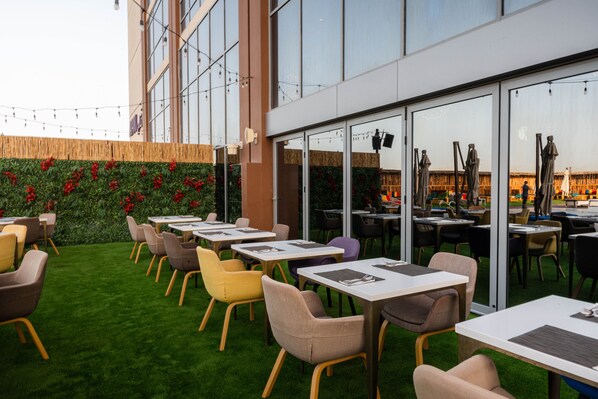 An outdoor seating area with tables and chairs, a green lawn, and a building with large windows. - Avani Ibn Battuta Dubai Hotel (Dubai)