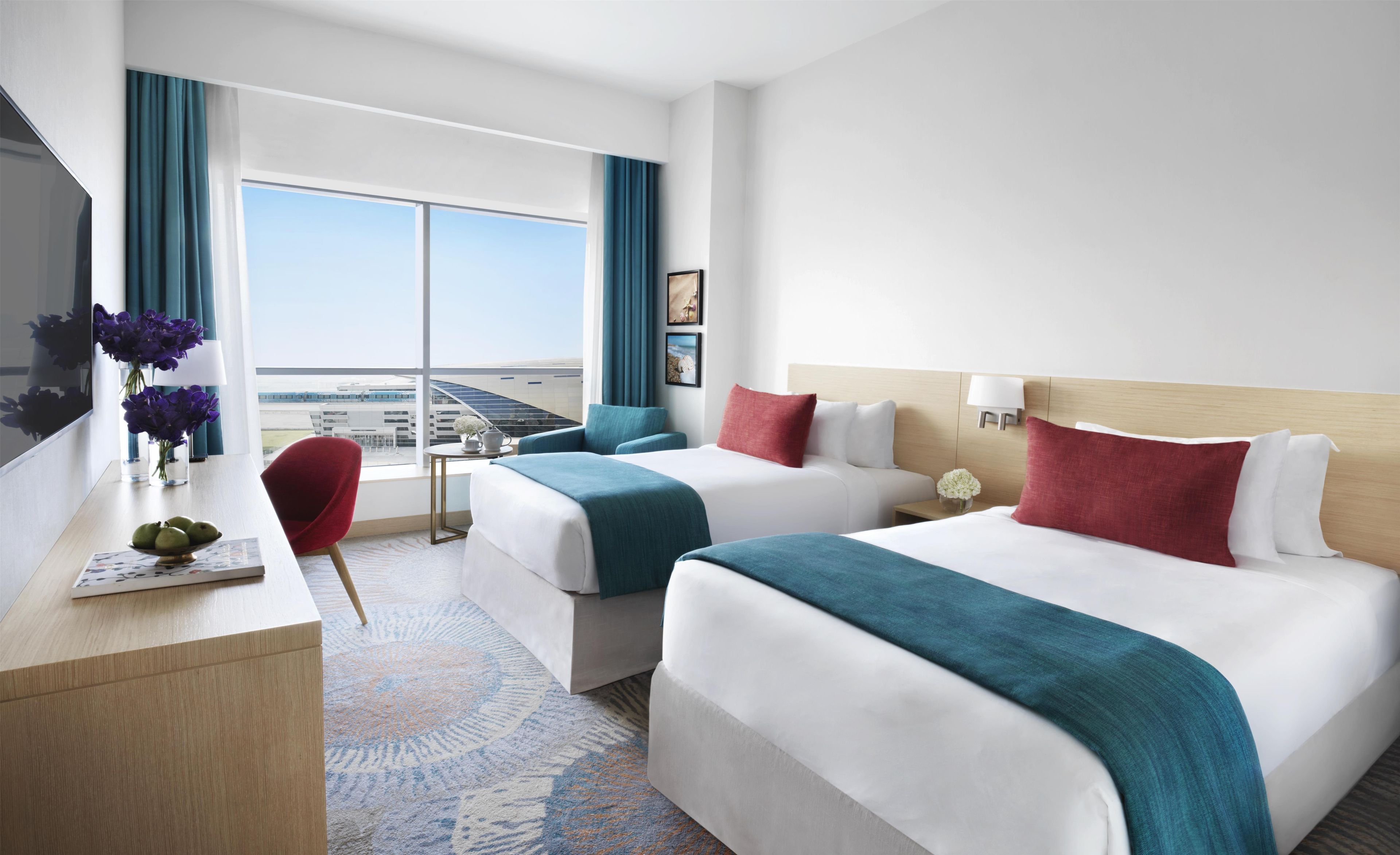 A hotel room with two beds, a desk, a TV, and a large window with a view of the airport.