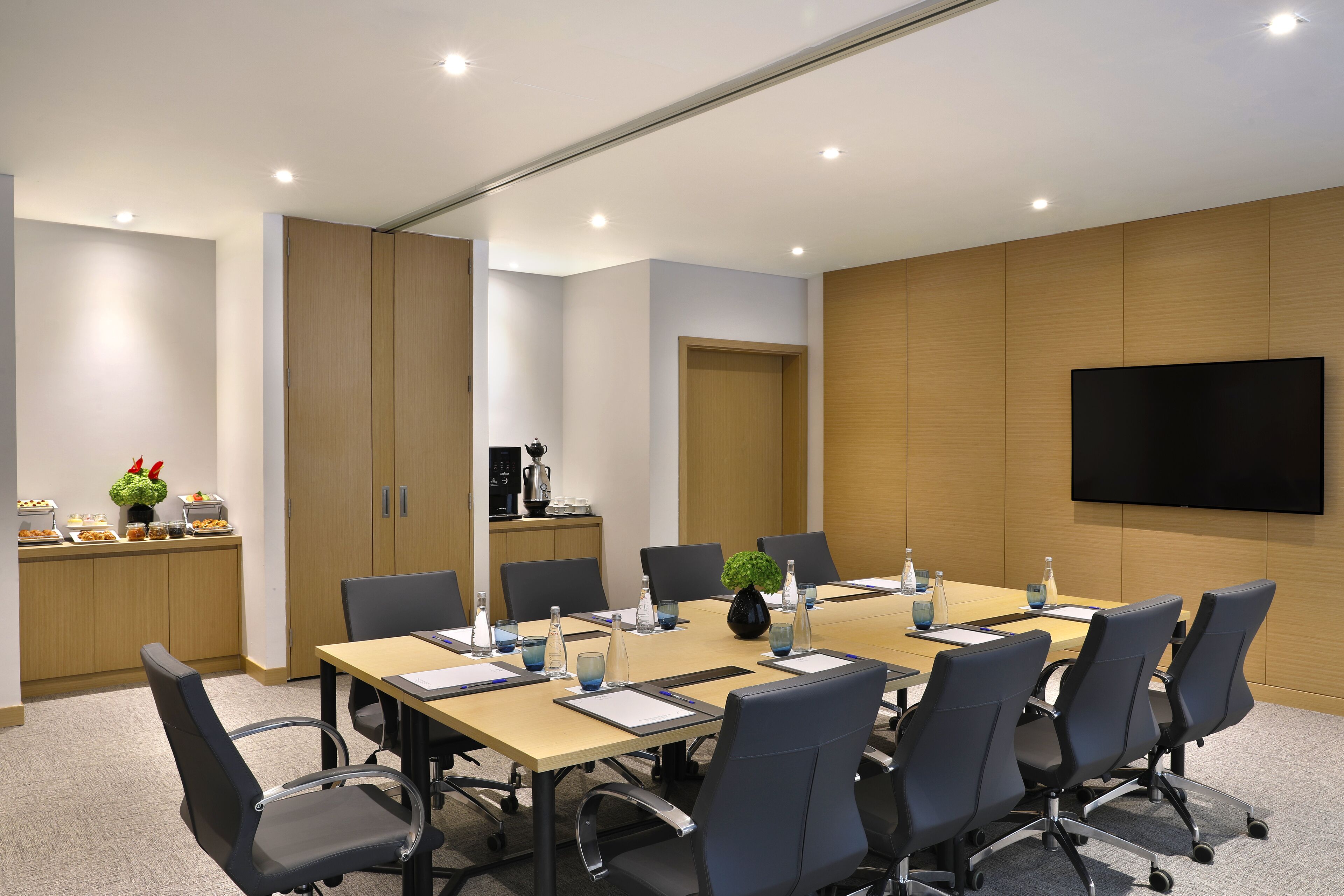 Sala de conferências com uma mesa comprida, cadeiras, uma TV e uma pequena copa.