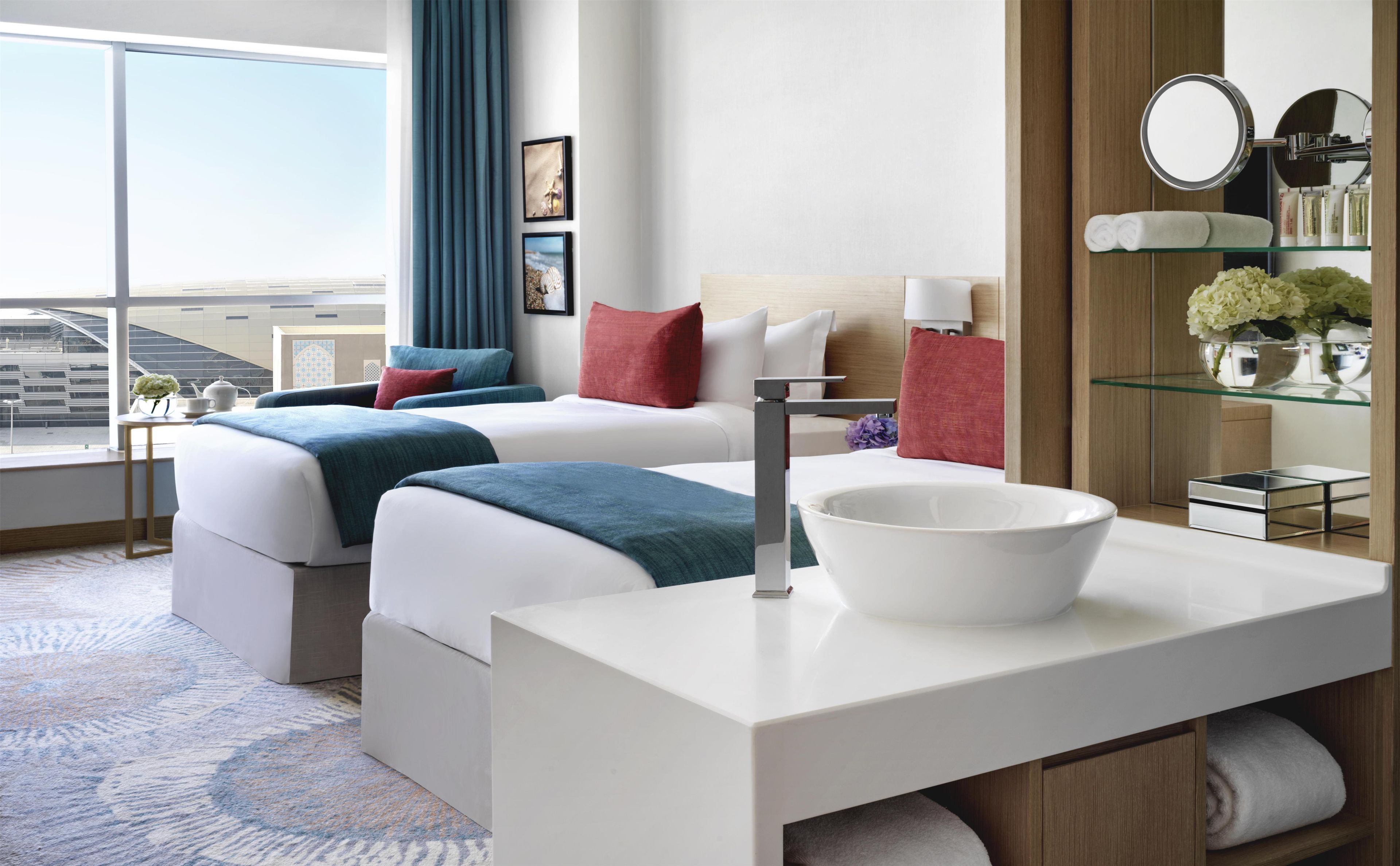 Une chambre d’hôtel avec deux lits, une grande fenêtre, une salle de bain moderne avec une étagère en verre et une vue sur le paysage urbain.