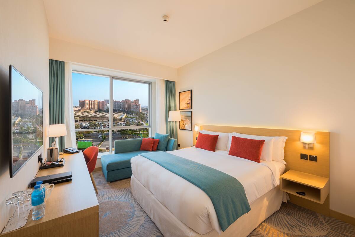 A hotel room with a large bed, a desk with a laptop, a chair, and a view of the cityscape.