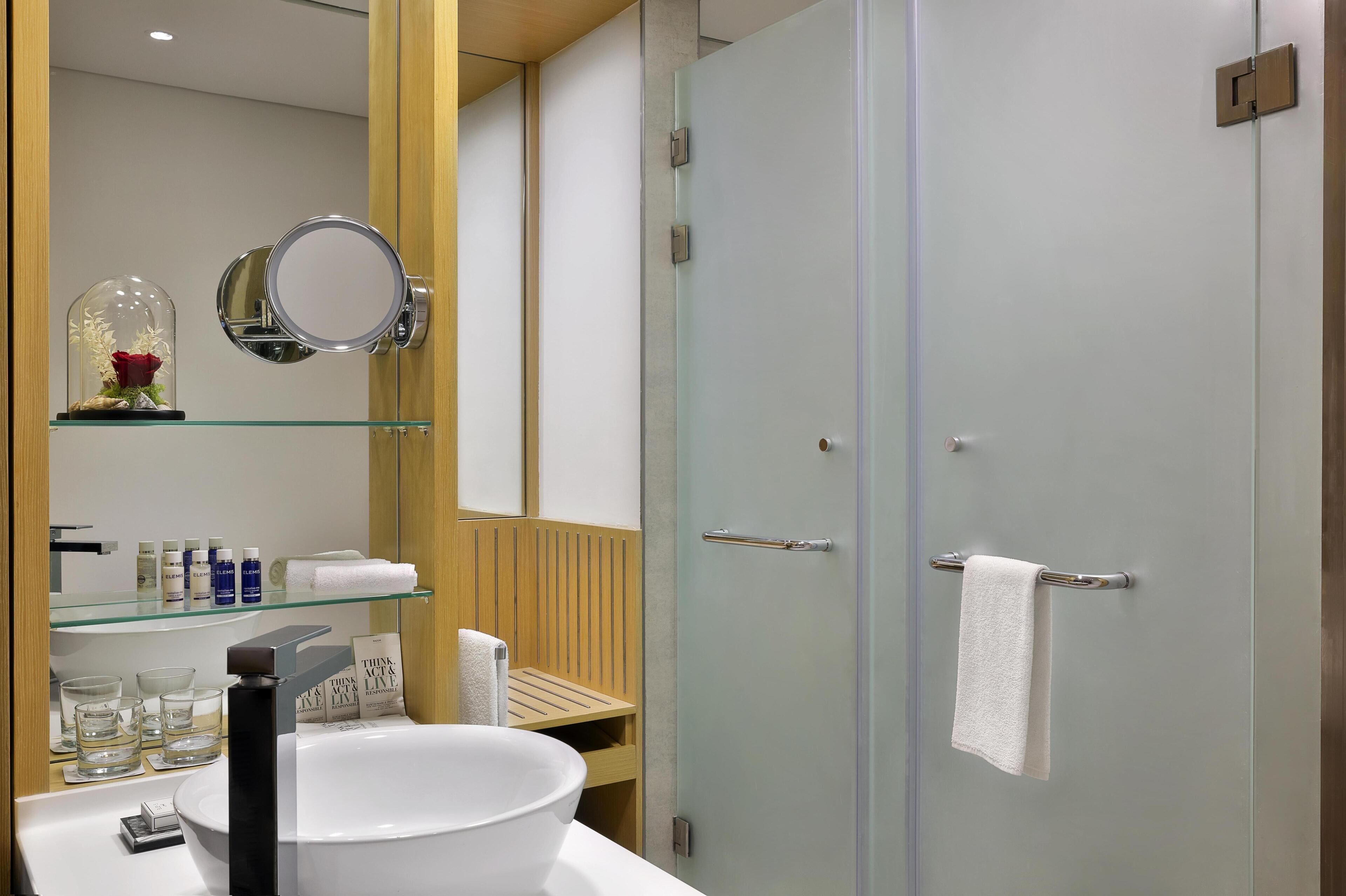 Ein modernes Badezimmer mit Duschkabine aus Glas, einem runden Spiegel und einem hölzernen Waschtisch mit Toilettenartikeln.