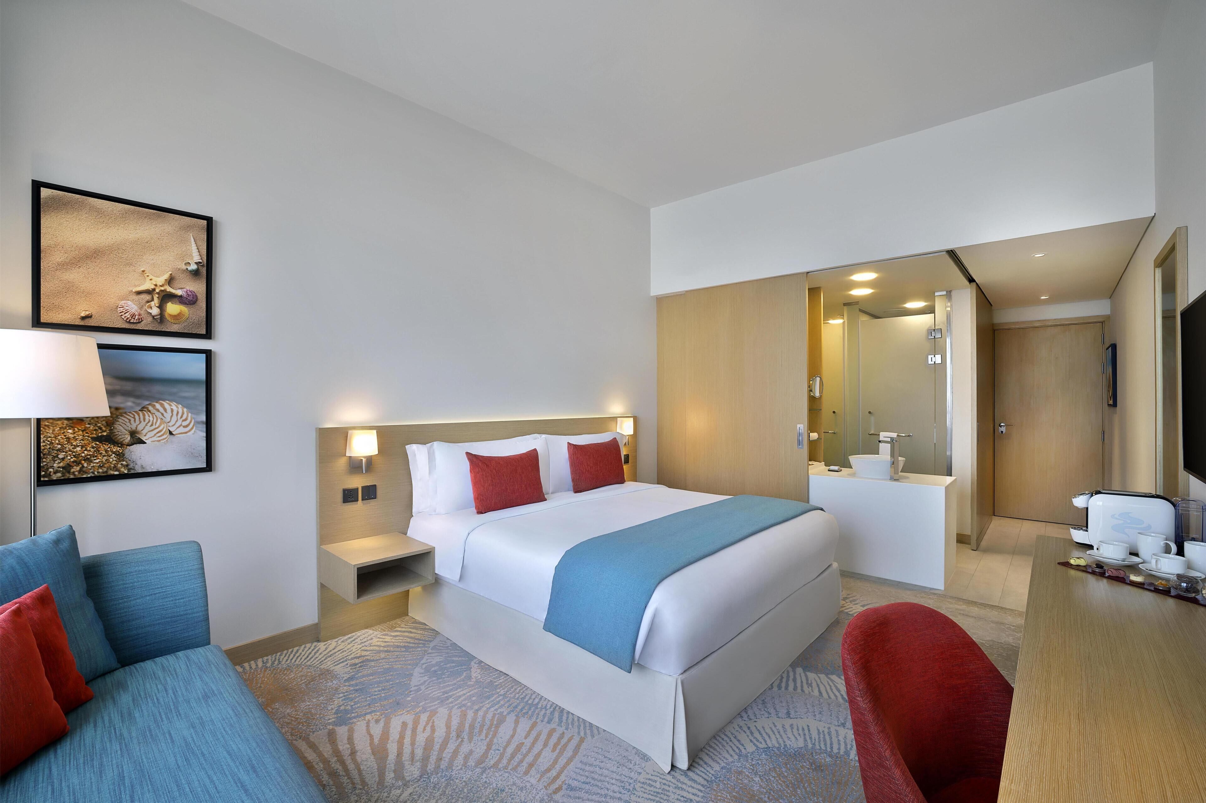 A modern hotel room with a large bed, a desk with a chair, a sofa, a bathroom, and a dining area.