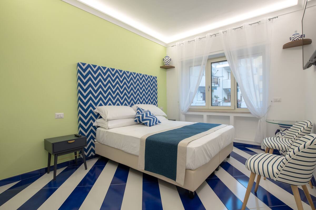 Un dormitorio con cama, mesita de noche, silla, ventana con cortina y pared con diseño de estampilla.