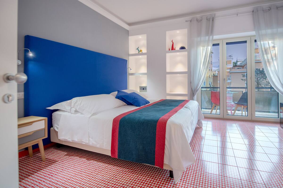 Un dormitorio con cama, mesita de noche, ventana con cortinas y vistas a edificios.