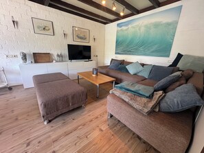 Een woonkamer met een bruine bank, een houten salontafel, een televisie op een witte kast, en een groot schilderij van een golf op de muur.