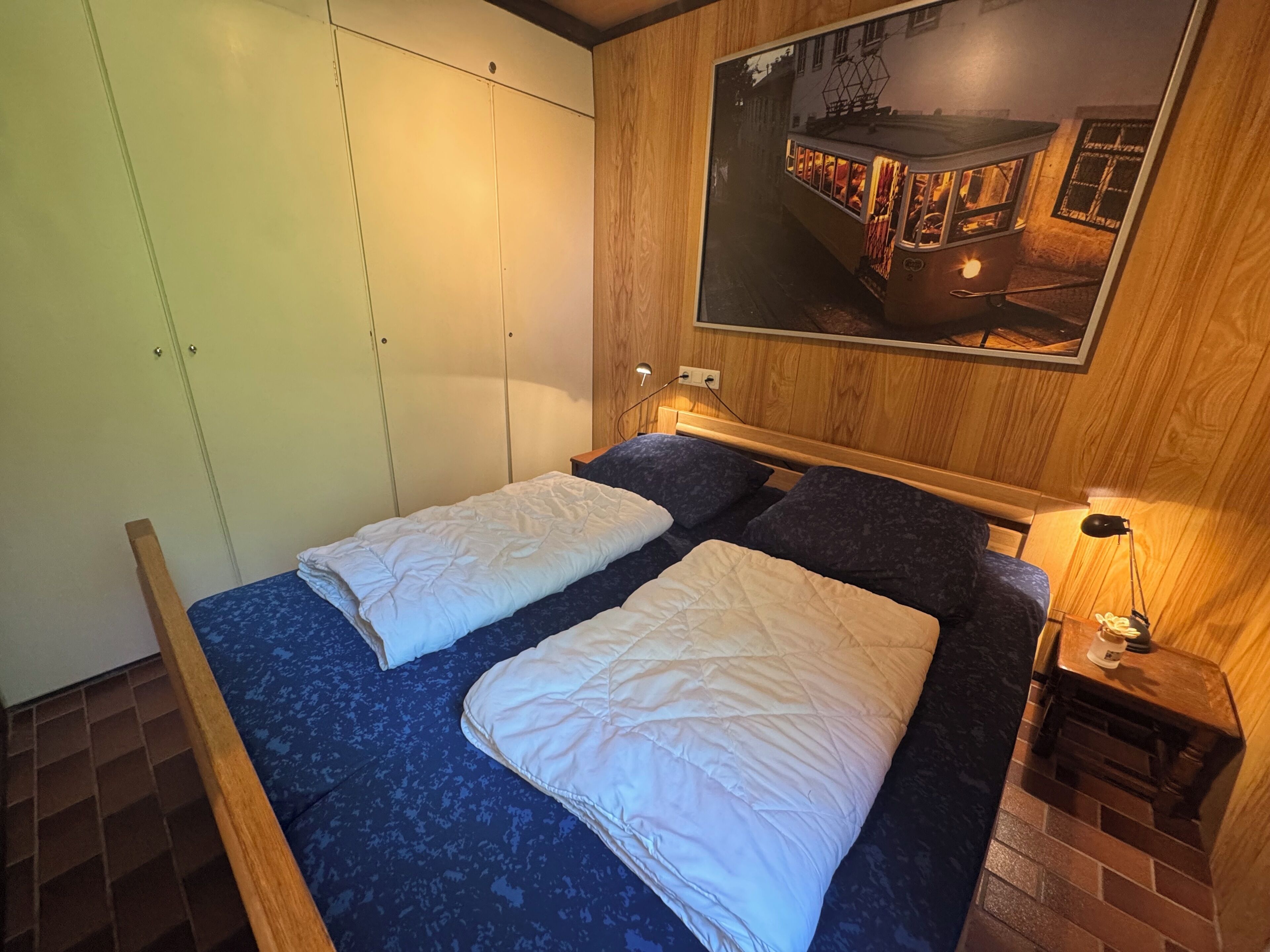 Una camera con due letti, un'armadio in legno, un comodino con una lampada e un quadro appeso al muro raffigurante un tram.