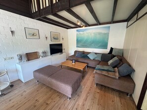 A living room with a white brick wall, wooden beams, a large painting, a flat-screen TV, a brown sofa, and a wooden coffee table.