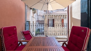 A wooden table with two red chairs and a beige umbrella in a balcony area with a view of a building with a tiled roof.